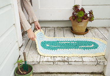 Two hands laying out a green, teal, and cream-colored rug on a whitewashed wooden porch.