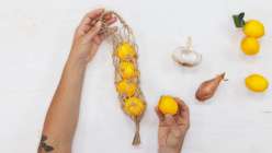 An overhead image of a hands holding a small twine bag containing lemons, aside a shallot and garlic from Cobrina's Knotted Pantry Bag Creativebug class.