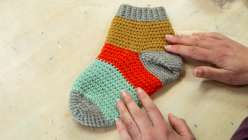 A crocheted sock in ochre, vermillion, and mint green yarn with two hands resting on top of it.