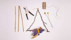 An image of a triangle of sticks with an assortment of dried flowers tied to the bottom alongside plant clippers, twine, three sticks and a pair of bright yellow billy button flowers.