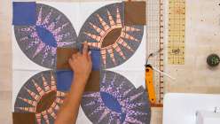 An overhead image of Sarah Bond pointing at a detail of her Pickle Wedges quilt block stitched in orange and grey fabrics, plus a rotary cutter and a quilting ruler.