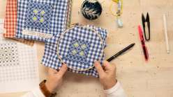 An overhead image of Blaire Stocker making chicken scratch embroidery, holding an embroidery hoop with navy blue gingham with stitching in blue and yellow.