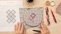 An overhead image of Blaire Stocker making chicken scratch embroidery, holding an embroidery hoop with taupe gingham with stitching in pink and red DMC embroidery thread.