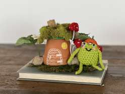 A happy crocheted frog next to a fairy house made using an upside-down terracotta pot from the Crafting Together: Mushroom Fairy Houses for Earth Day with Courtney and Twinkie Chan on Creativebug