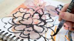 A hand holding a brush making an ink drawing of a flower over a collaged scrapbook page from Courtney Cerruti's Creativebug class Find Your Creative Rhythm: 15 Ways to Spark & Sustain Art Making.