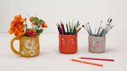 A grey and orange vase, both with pink cheeks and skeptical expressions, and a yellow pitcher with flower details made by Cindy Willingham in her Hand-Built Vases with Air-Dry Clay class on Creativebug.