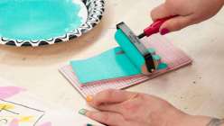 Risa Iwisaki Culbertson using a brayer to roll out teal-colored acrylic paint onto a hand-carved stamp pad, made in her class Wearable Wins on Creativebug