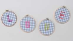 Image of four embroidery hoops on a table, stretched with blue gingham and letters cross-stitched to spell 